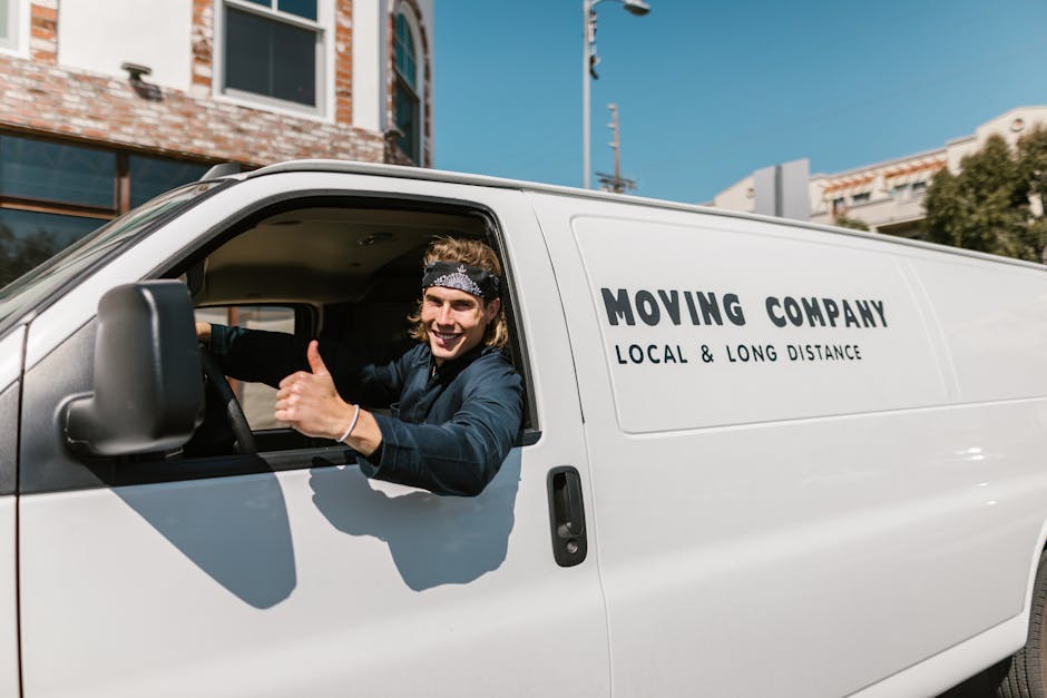 A young man with long, wavy hair, wearing a patterned headband and a dark blue shirt, is sitting in the driver's seat of a white moving van with the words 'MOVING COMPANY' and 'LOCAL & LONG DISTANCE' 