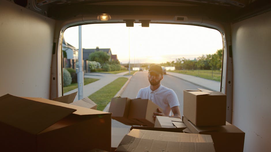 A young man with long, wavy hair, wearing a patterned headband and a dark blue shirt, is sitting in the driver's seat of a white moving van with the words 'MOVING COMPANY' and 'LOCAL & LONG DISTANCE' 