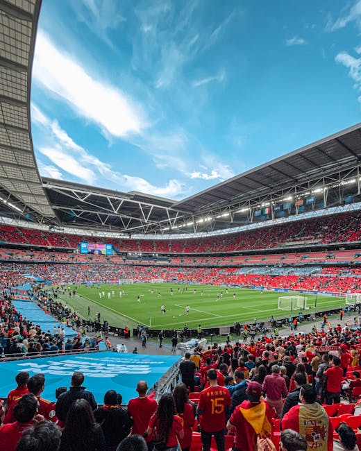 A panoramic view of Twickenham Stadium during a live sporting event, with a large audience seated in the tiered red and gray stands surrounding the lush green pitch. The stadium's roof structure extends over the seating areas, and a bright blue sky with scattered white clouds is visible overhead. The field is occupied by players engaged in a match, with some visible near the goals and others positioning across the pitch. In the foreground, spectators are seated and standing along the lower levels, dressed in various colors, some wearing team jerseys. The overall scene captures the atmosphere of a major sporting occasion, suitable for contextual relevance to home relocation and transport logistics, aligning with professional moving services such as those offered by Man with Van Twickenham, particularly in relation to relocating or transporting sports facilities or equipment.