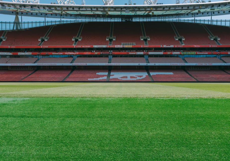 A view of the interior of Twickenham Stadium showing the pitch with neatly mowed grass in varying shades of green, from light to dark, indicating the area designated for home relocation activities. Behind the pitch, there are rows of red stadium seating arranged in a tiered formation with some sections labelled with the years 1994, 1998, 2002, and 2004. The seating area features a large letter 'T' visible in the middle section, spelled out with white seats amidst the red ones. Above the seating, the stadium's roof structure extends outward, supported by metal beams and covered with a translucent material, allowing natural light to illuminate the interior. The upper levels of the stadium display banners and advertising displays, including a visible URL for Arsenal.com, indicating ongoing sports or event preparations. This environment offers a clear perspective of the stadium's seating and field area, relevant for illustrating professional removal or transport logistics associated with events at Twickenham, as documented by Man with Van Twickenham.