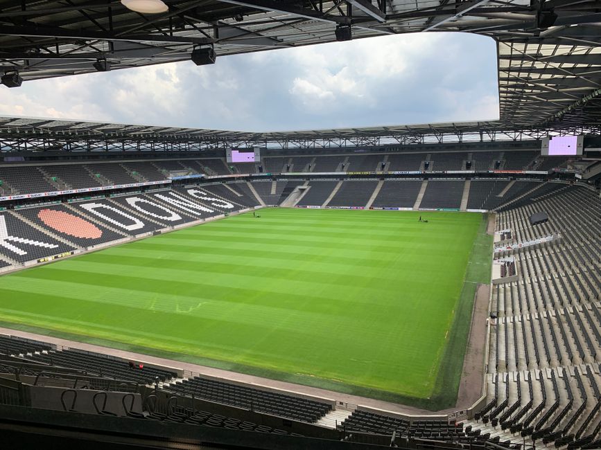 A panoramic view of Twickenham Stadium during a live sporting event, with a large audience seated in the tiered red and gray stands surrounding the lush green pitch. The stadium's roof structure extends over the seating areas, and a bright blue sky with scattered white clouds is visible overhead. The field is occupied by players engaged in a match, with some visible near the goals and others positioning across the pitch. In the foreground, spectators are seated and standing along the lower levels, dressed in various colors, some wearing team jerseys. The overall scene captures the atmosphere of a major sporting occasion, suitable for contextual relevance to home relocation and transport logistics, aligning with professional moving services such as those offered by Man with Van Twickenham, particularly in relation to relocating or transporting sports facilities or equipment.